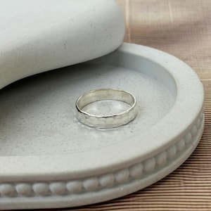 May include: A silver ring with a hammered texture sits on a light gray, round, decorative tray. The ring has a simple band design. The tray has a beaded edge and is partially covered by a curved, light gray lid.