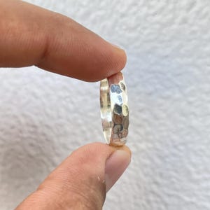 May include: A hammered silver ring with a textured surface. The ring is held between two fingers, showcasing its reflective qualities. The ring's design features a series of small, irregular indentations, creating a unique, eye-catching appearance.