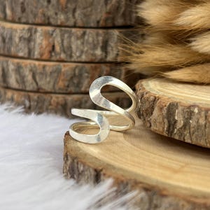 May include: A silver-toned ring with an open, abstract design. The ring features a curved, flowing shape with an open oval at the top and a smaller, curved element below. The ring is displayed on a wooden surface.