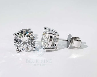 May include: A pair of diamond stud earrings with a brilliant cut. The earrings are set in a silver metal, with a post and butterfly backing. The earrings are displayed on a white surface. The brand name "BLUE FINE JEWELS" is visible.
