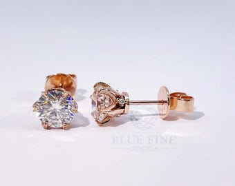 May include: Rose gold stud earrings with a clear, round gemstone. The earrings have a decorative filigree design around the gemstone. The image also includes the text "BLUE FINE JEWELS".