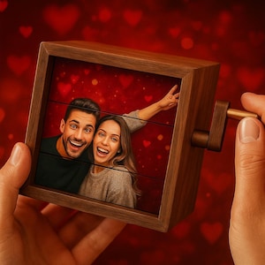 Könnte beinhalten: Ein hölzerner Fotowürfel mit Kurbel, der ein lächelndes Paar vor einem roten Hintergrund mit Herzformen zeigt. Der Rahmen des Würfels ist warmbraun, und die Kurbel wird von einer Hand gehalten.