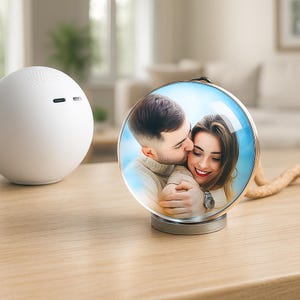 Può includere: Una cornice rotonda color argento con una cupola di vetro trasparente, che mostra una coppia che si abbraccia e si bacia. La cornice è su una superficie di legno, con un altoparlante sferico bianco a sinistra. Una corda beige è attaccata alla cornice.