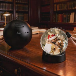 Op de afbeelding: Een zwarte, bolvormige luidspreker met een USB-poort en een heldere sneeuwbol met een kussend stel onder een kerstmuts. De sneeuwbol staat op een zwarte voet en heeft een zwart-roze koord.