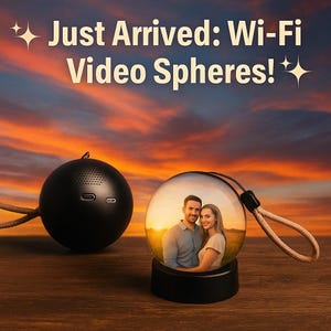 May include: The image shows two black video spheres. One is a speaker, and the other is a clear sphere with a couple inside. The text "Just Arrived: Wi-Fi Video Spheres!" is at the top. The spheres are on a wooden surface against a sunset background.