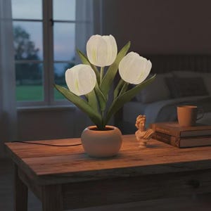 May include: A white tulip-shaped lamp with three illuminated flower heads and green leaves, set in a small, round, beige pot. The lamp sits on a wooden table, with a small statue and books nearby.