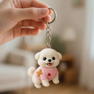 May include: A white puppy keychain with a pink sweater featuring a yellow star and a red apple. The keychain has a silver chain and ring. The puppy has black eyes and a pink tongue.