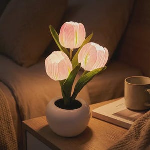 May include: A decorative tulip night light with three pink and white illuminated flower heads and green leaves. The tulips are in a small white ceramic pot, sitting on a wooden nightstand. A mug and book are also on the nightstand.