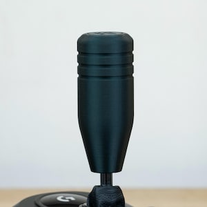 Może przedstawiać: Czarny, teksturowany gałka zmiany biegów z cylindrycznym wierzchem i zwężającym się korpusem. Gałka jest zamontowana na czarnym wałku i jest częścią kontrolera do gier. Tło jest gładkie białe.