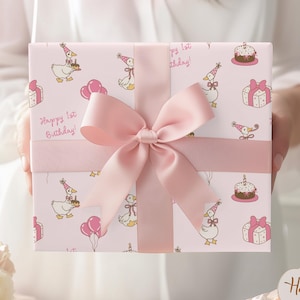 May include: A wrapped gift box with a pink bow. The wrapping paper is light pink with a pattern of cartoon geese wearing party hats, cakes, balloons, and gift boxes. The text "Happy 1st Birthday!" is printed on the wrapping paper.