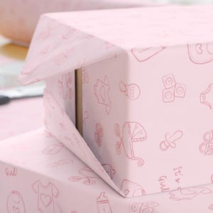 K&ouml;nnte beinhalten: Rosa Geschenkbox, verpackt in Papier mit einem sich wiederholenden Muster von Baby-Illustrationen in einem helleren Rosa. Die Illustrationen umfassen eine Babyflasche, einen Strampler, einen Schnuller und einen Kinderwagen. Die Worte "It's a Girl" sind sichtbar.
