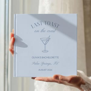 May include: A square, light blue photo album with dark blue text and a martini glass graphic. The cover reads: LAST TOAST on the coast, OLIVIA'S BACHELORETTE, Palm Springs, FL, AUGUST 2026. The design features a centered cocktail glass illustration.