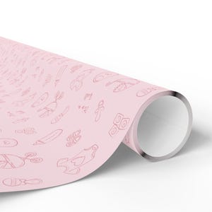 K&ouml;nnte beinhalten: Eine Rolle rosa Geschenkpapier mit einem sich wiederholenden Muster von Baby-Themen-Kritzeleien in dunklerem Rosa. Die Kritzeleien umfassen Schnuller, Babyschuhe und andere Baby-Artikel. Der Text "It's a girl" ist ebenfalls sichtbar.