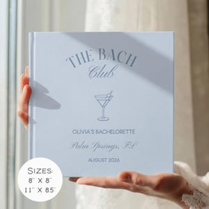 May include: A light blue square hardcover book with dark blue text and a martini glass graphic. The cover reads THE BACH CLUB, OLIVIA'S BACHELORETTE, Palm Springs, FL, AUGUST 2026. A white circular label indicates available sizes of 8 inches by 8 inches.