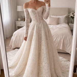 May include: A floor-length off-the-shoulder wedding dress featuring long sleeves and a full, voluminous skirt. The entire gown is crafted from white lace with a detailed floral pattern, a sweetheart neckline, and a structured bodice.