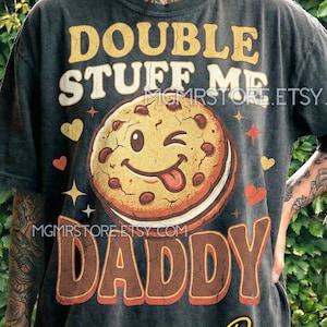 Peut inclure: T-shirt gris foncé avec l'inscription "DOUBLE STUFF ME DADDY" dans une typographie rétro. Un dessin de biscuit souriant avec des pépites de chocolat et une langue est au centre. Des cœurs et des étoiles sont également présents.