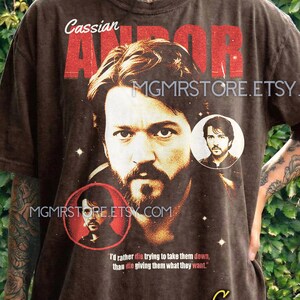 May include: Espresso-colored t-shirt featuring a portrait of a man with the text "Cassian ANDOR" in red. The shirt includes two smaller circular portraits and the quote, "I'd rather die trying to take them down, than die giving them what they want."