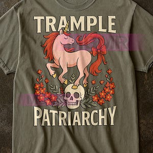 May include: Olive green t-shirt featuring a pink unicorn with a red mane and tail, standing on a skull. The text "TRAMPLE PATRIARCHY" is printed above and below the unicorn graphic. The design includes floral elements.