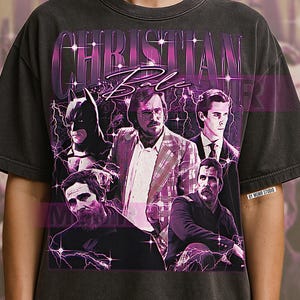 Può includere: T-shirt grigio scuro con una grafica di Christian Bale e la scritta "CHRISTIAN BALE". Il design include immagini di Bale in vari ruoli, con un motivo a fulmini viola e bianchi.
