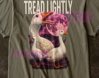 Op de afbeelding: Olijfgroen t-shirt met een afbeelding van drie ganzen voor een roze maan. De tekst "TREAD LIGHTLY" staat in witte blokletters bovenaan. De ganzen hebben witte veren en oranje snavels.