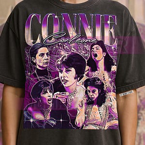 May include: Dark gray t-shirt featuring a vintage-style graphic of the name "CONNIE" above a collage of women's faces. The design has a purple and white color scheme with lightning bolt accents, creating a retro aesthetic.