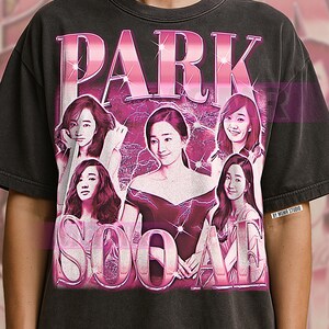 Puede incluir: Camiseta gris oscuro con un gráfico rosa que presenta el nombre "PARK SOO AME" e imágenes de una mujer. El diseño tiene un aspecto vintage y desgastado. La camiseta es de cuello redondo y tiene mangas cortas.