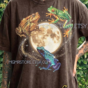 Può includere: T-shirt marrone con una grafica di tre rane colorate davanti a una luna piena e stelle. La maglietta ha il testo "MGMSTORE.ETSY.COM" e "MGMSTORE" stampato sopra. La maglietta ha anche la parola "Espresso" stampata sopra.