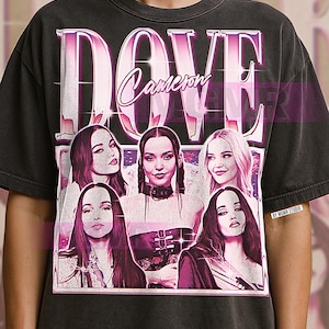 May include: Dark gray t-shirt featuring a graphic of Dove Cameron with the name "DOVE" in large, pink, retro-style lettering above her name. The shirt has a vintage aesthetic.