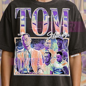 May include: Dark gray t-shirt featuring a graphic of Tom Hanks with the name "TOM Hanks" in large, stylized letters. The graphic includes multiple images of the actor in various roles, with a vintage, distressed aesthetic.