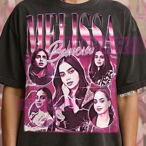 May include: Dark gray t-shirt featuring a graphic design with the name "MELISSA BARRERA" in a pink, retro-style font. The design includes multiple images of a woman, with a pink lightning bolt effect. The shirt has a vintage look.