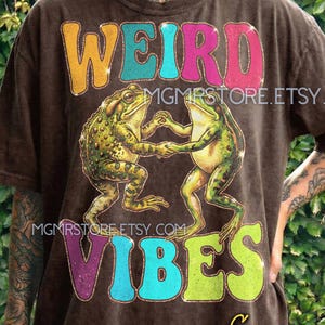 May include: Brown t-shirt with the words "WEIRD VIBES" in colorful, glittery letters. Two green frogs are dancing below the text. The shirt also features the Etsy shop name "MGMRSTORE.ETSY.COM".