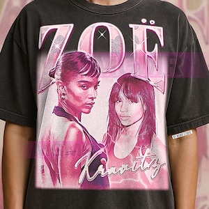 Op de afbeelding: Donkergrijze t-shirt met een roze afbeelding van twee vrouwen, de tekst "ZOE" erboven en "Kravitz" eronder. Het ontwerp heeft een vintage, vervaagde uitstraling, met een roze en wit kleurenschema.