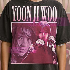 May include: Dark gray t-shirt featuring a graphic of a woman with the text "YOON JI WOO" in a pink font. The image has a vintage, distressed look. The t-shirt is a crew neck style.
