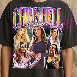 May include: Dark gray t-shirt featuring a graphic design with the text "Chrishell Stause" in a retro style. The design includes images of several women. The shirt has a vintage, worn look.