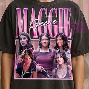 May include: Dark gray t-shirt featuring a pink and purple graphic with the name "Maggie Greene" and multiple images of a woman. The design has a vintage, distressed look.