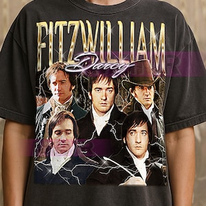 May include: Dark gray t-shirt featuring a vintage-style graphic of Fitzwilliam Darcy with the text "Fitzwilliam Darcy" in gold lettering. The graphic includes portraits of actors. The shirt has a classic crew neck.