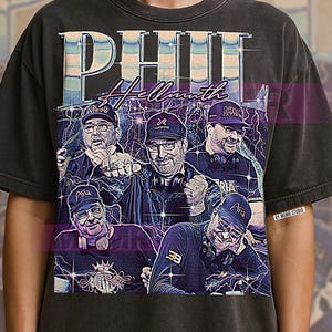 Pode incluir: T-shirt cinzento escuro com um gráfico retro de Phil Hellmuth e o texto "Phil Hellmuth". O gráfico inclui várias imagens de Phil Hellmuth com detalhes de raios. A t-shirt tem um visual vintage.