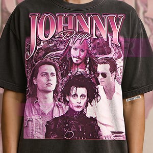 Könnte beinhalten: Ein anthrazitfarbenes Kurzarm-T-Shirt mit einem großen, rosafarbenen Grafikdruck von Johnny Depp in verschiedenen Filmrollen. Der Text "JOHNNY" erscheint in großen rosa Buchstaben oben, darunter "Depp". Ein kleines "BY MGMR STUDIO"-Etikett ist auf der rechten Seite zu sehen.