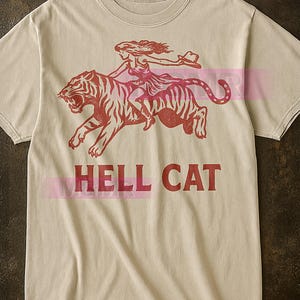 Könnte beinhalten: Beiges T-Shirt mit roter Grafik einer Frau, die auf einem Tiger reitet und einen Hut hält. Der Text "HELL CAT" ist darunter in Rot gedruckt. Das Shirt hat einen Rundhalsausschnitt und kurze Ärmel.