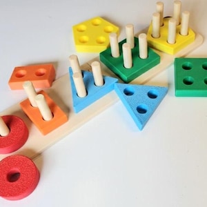 May include: A colorful wooden shape sorter toy with various geometric shapes, including circles, squares, triangles, and a pentagon. The shapes are in red, orange, yellow, green, and blue, with wooden pegs for matching. The toy is designed for early childhood development.