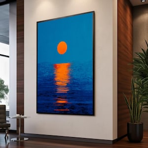 Könnte beinhalten: Ein gerahmter Kunstdruck zeigt einen Sonnenuntergang über dem Wasser. Der Himmel ist tiefblau, mit einer leuchtend orangefarbenen Sonne. Das Wasser reflektiert das Sonnenlicht und erzeugt einen schimmernden Effekt. Das Kunstwerk ist in einem schwarzen Rahmen.