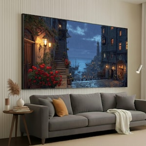 May include: A large framed canvas print of a European coastal town at dusk. The artwork shows a cobblestone street, buildings with lit windows, and a distant lighthouse. Red flowers and a staircase are visible on the left. The scene is set against a dark, cloudy sky.