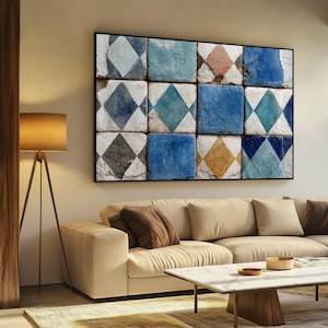 Op de afbeelding: Een ingelijst kunstwerk met een raster van vierkante tegels met ruitpatronen in blauw, grijs en geel. Het kunstwerk hangt boven een beige bank in een woonkamer.