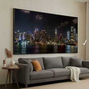 Puede incluir: Un gran lienzo enmarcado que representa un paisaje urbano nocturno, con rascacielos iluminados que se reflejan en el agua. La impresión cuelga sobre un sofá gris con cojines decorativos y una manta.