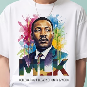 May include: White t-shirt featuring a portrait of Martin Luther King Jr. with the letters "MLK" in bold, colorful text. The design includes a vibrant watercolor splash background and the text "CELEBRATING A LEGACY OF UNITY & VISION".
