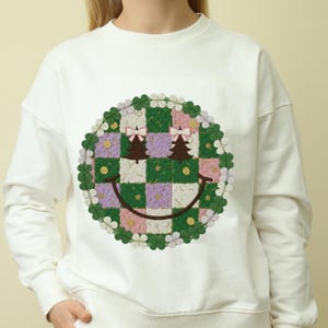 May include: White sweatshirt featuring a large, embroidered smiley face design. The smiley face is composed of a checkered pattern in green, purple, and pink, with two Christmas trees and bows. The design is surrounded by a floral border.