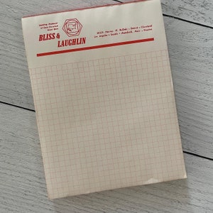 Op de afbeelding: Een vintage notitieblok met een rood-witte koptekst met de tekst "BLISS & LAUGHLIN" en "Leading Producer of Cold Finished Steel Bars". Het notitieblok heeft een rasterpatroon op de pagina's.