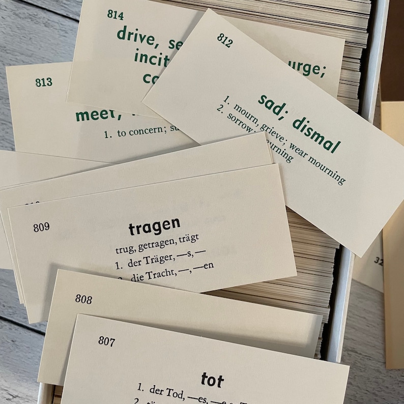 May include: A collection of flashcards with green text and numbers. The cards are white and feature vocabulary words and definitions. Some cards are labeled with German words like "tragen" and "tot". The cards are stacked in a box.