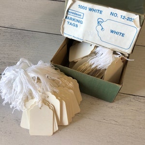 May include: A box of white Dennison marking tags, with a scalloped edge design, and white string attached. The box is open, revealing a stack of tags. The box is green and white, with the text "1000 WHITE" and "MARKING TAGS".
