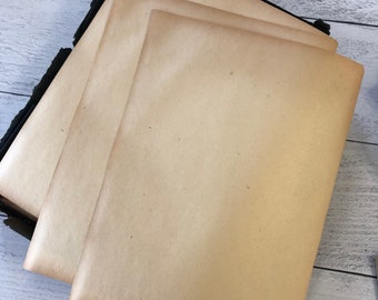 May include: A stack of aged, tan-colored paper sheets. The paper has a slightly textured surface and a warm, vintage hue. The edges of the paper are visible, and the stack is held together by a black binding. The background is a light-colored, wooden surface.